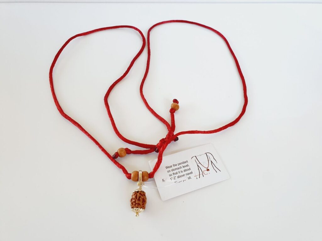 Red string necklace with a 3 mukhi Rudraksha pendant in silver displayed on a white surface.