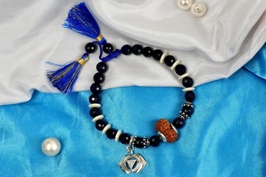 Spiral bracelet with 14 Mukhi Rudraksha beads, blue sapphire gemstone beads, Third-eye chakra pendant, wooden and Lava stone beads on white and blue fabric