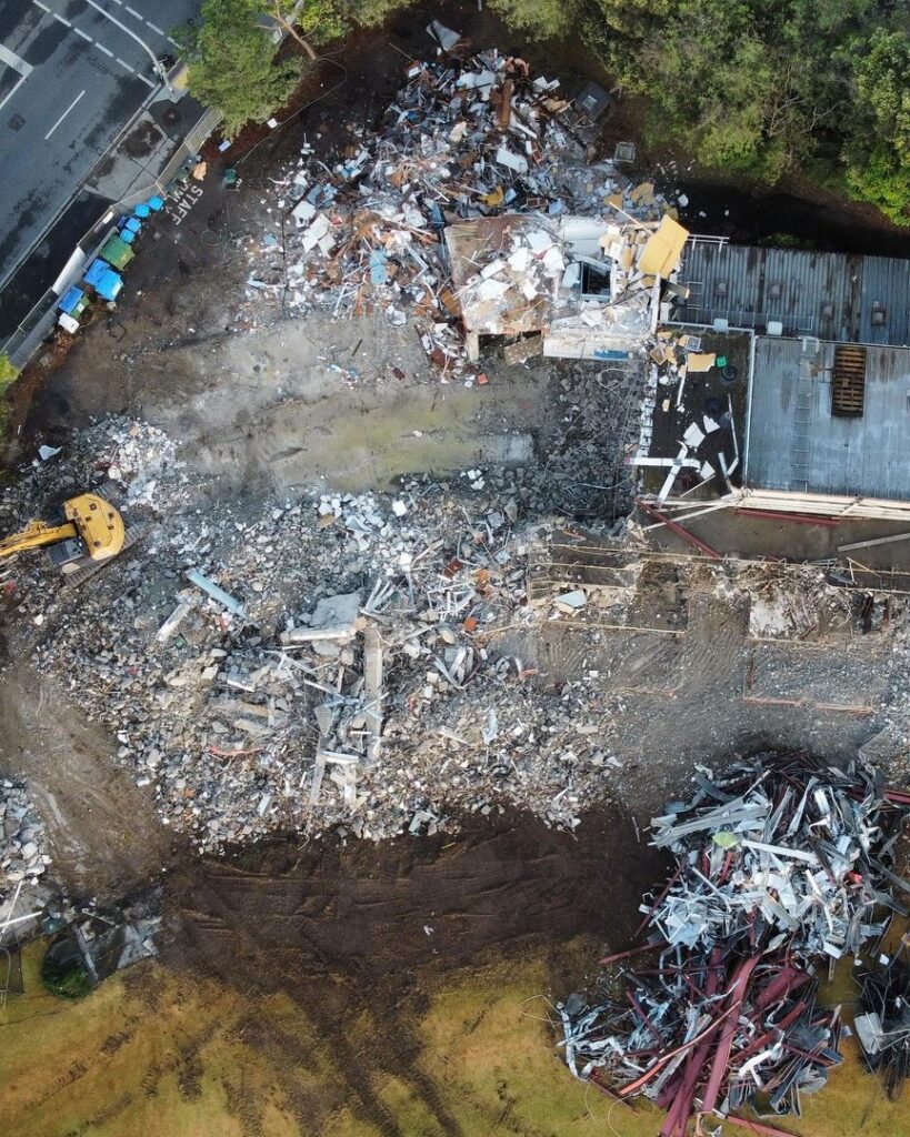 Aerial view of a demolition site with an excavator, rubble, and partially demolished buildings.