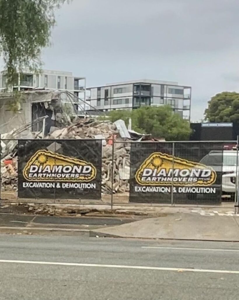 Demolition site with Diamond Earthmovers banners, rubble, and modern apartments in the background.