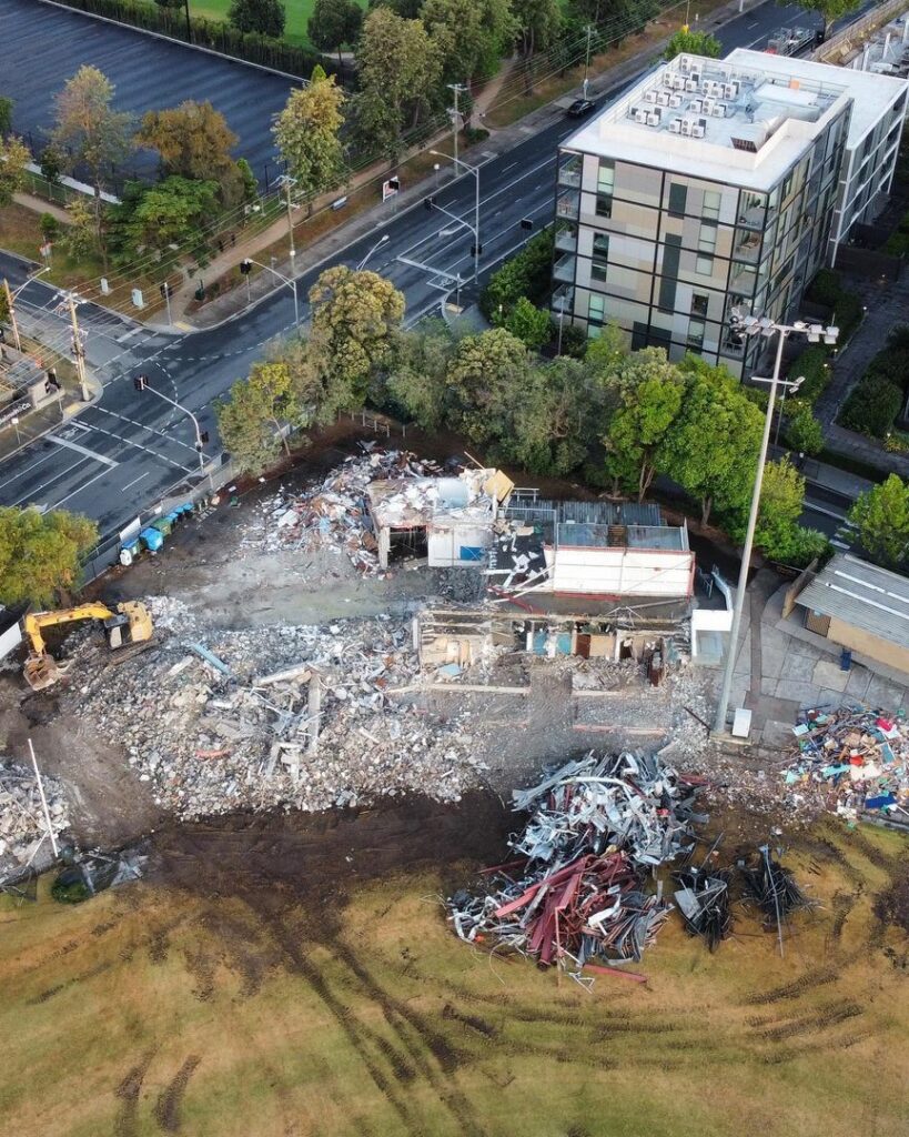 Aerial view of a demolition site with debris, an excavator, and a partially demolished building near urban roads.