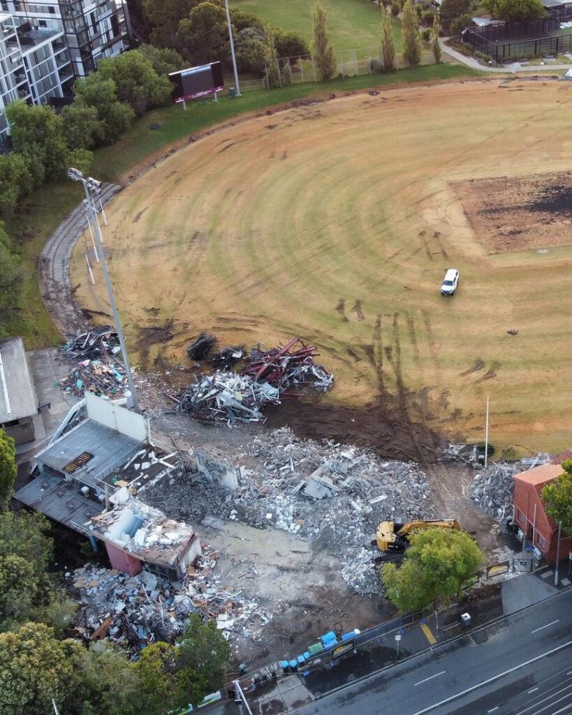 Excavator demolishing grandstand club rooms at an outdoor sports field with visible debris and surrounding environment.