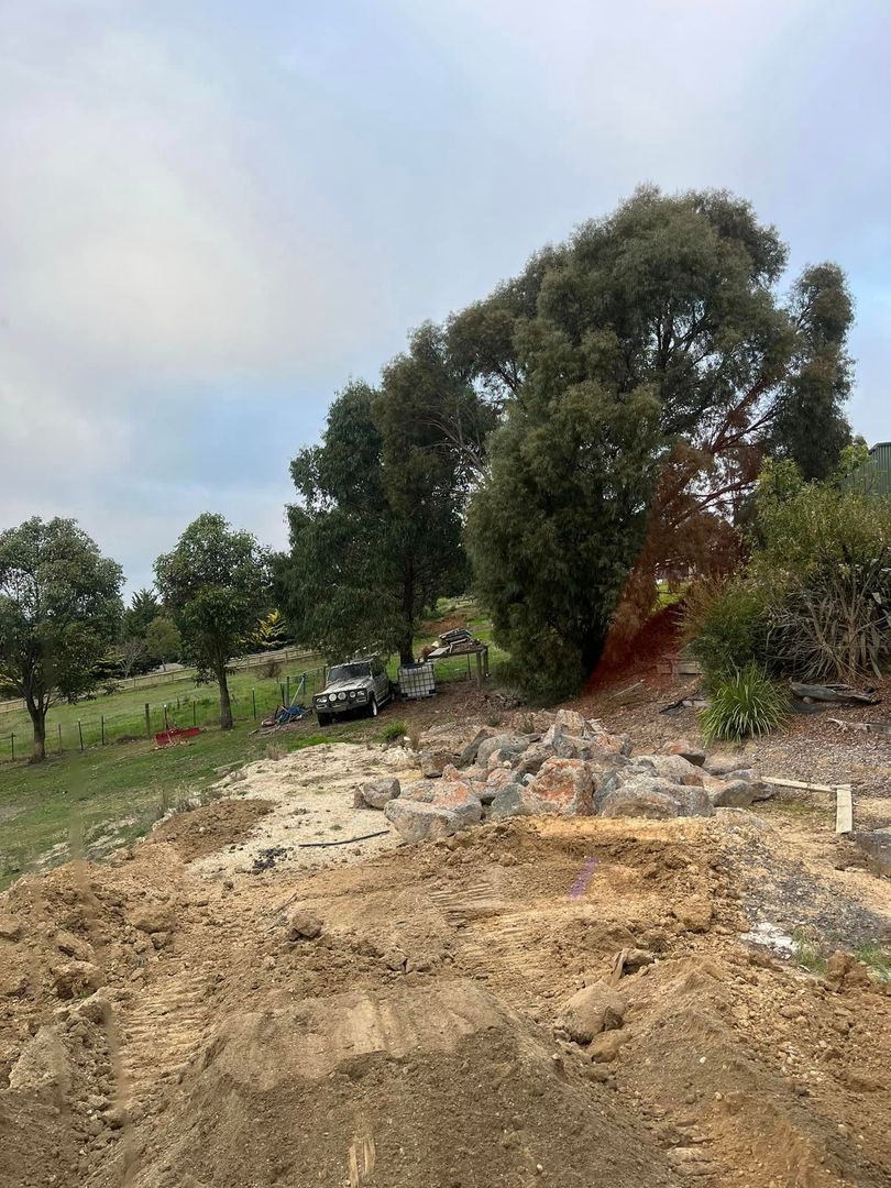 Excavation site with dirt, basalt rocks, a parked vehicle, and trees in a rural setting.