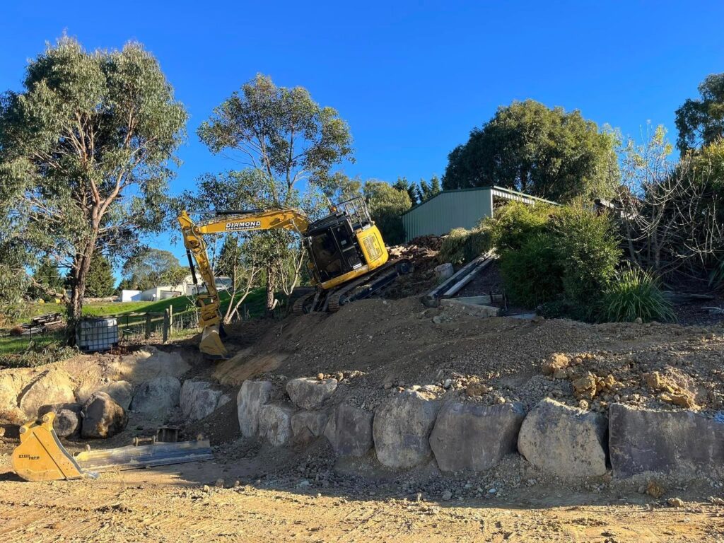 Yellow excavator constructing a basalt rock retaining wall at an outdoor site with trees and a green shed in the background.