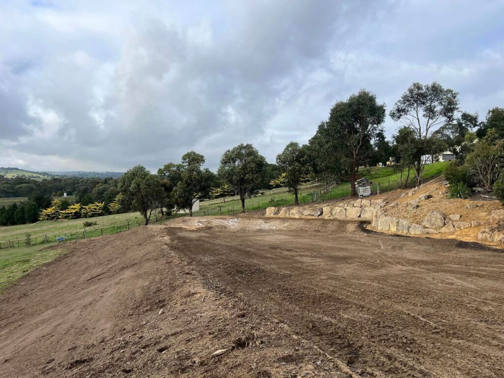 A cleared dirt pad with a basalt rock retaining wall in a rural area, surrounded by grassy fields and trees under an overcast sky.