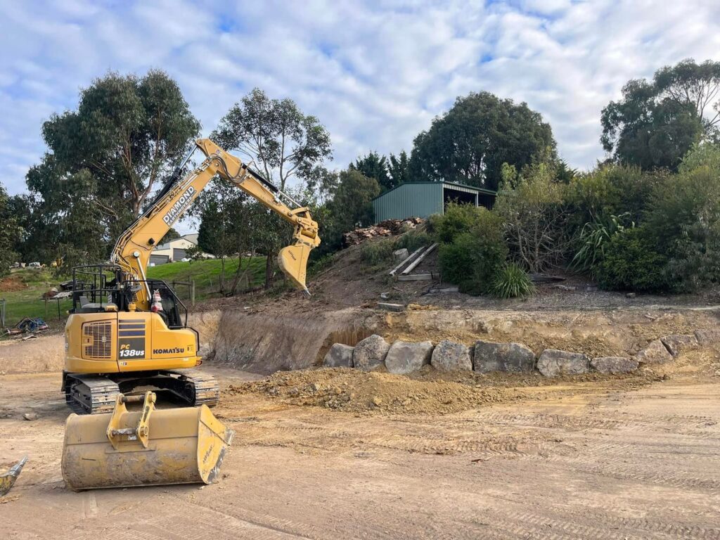 Yellow Komatsu PC138US excavator working on a dirt area with a basalt rock retaining wall and green shed in the background.