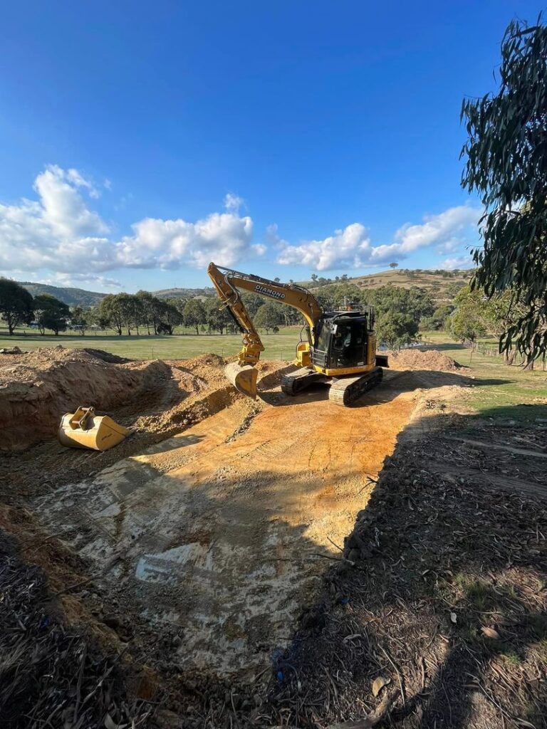 A yellow Diamond excavator on a leveled dirt pad in a rural field with trees and hills in the background.