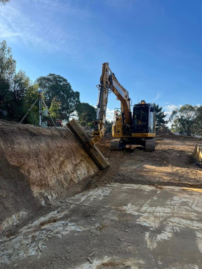 An excavator using 3D GPS technology works on a dirt slope at an outdoor construction site with trees in the background.