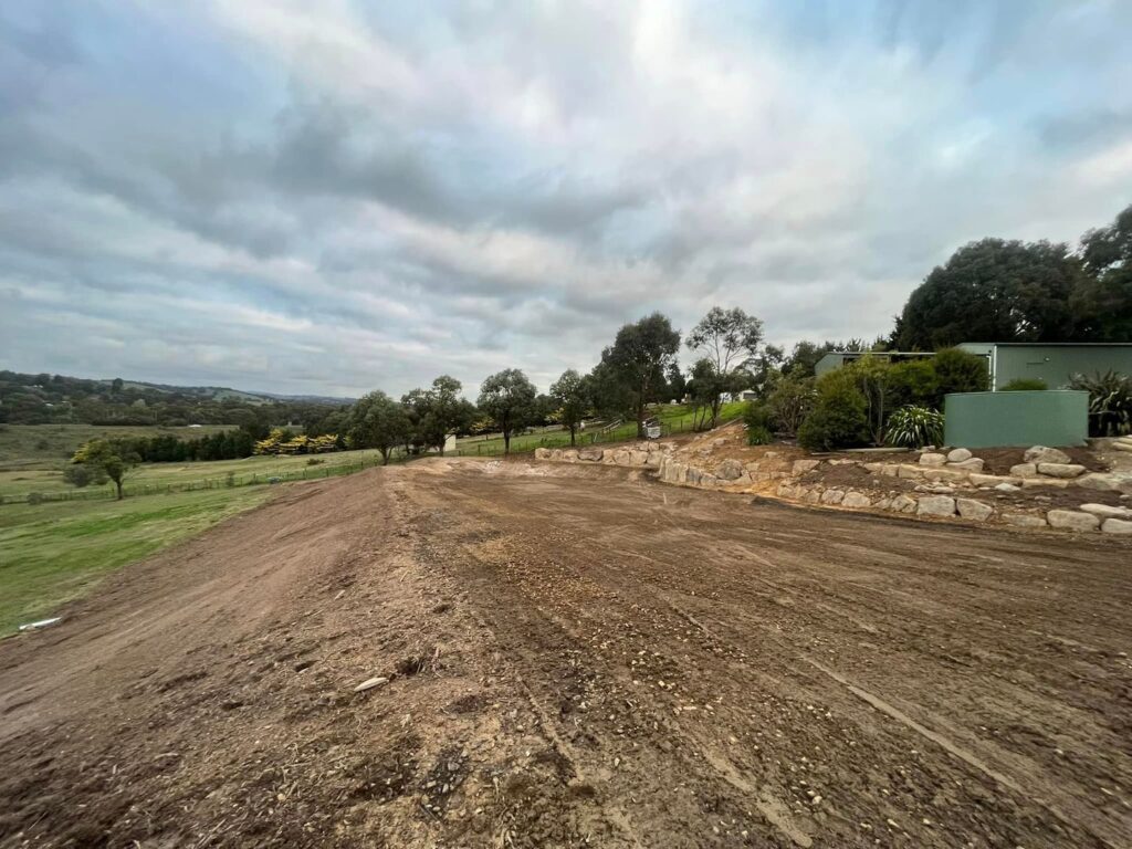 Cleared dirt pad with basalt rock retaining wall and green structure in a rural setting.