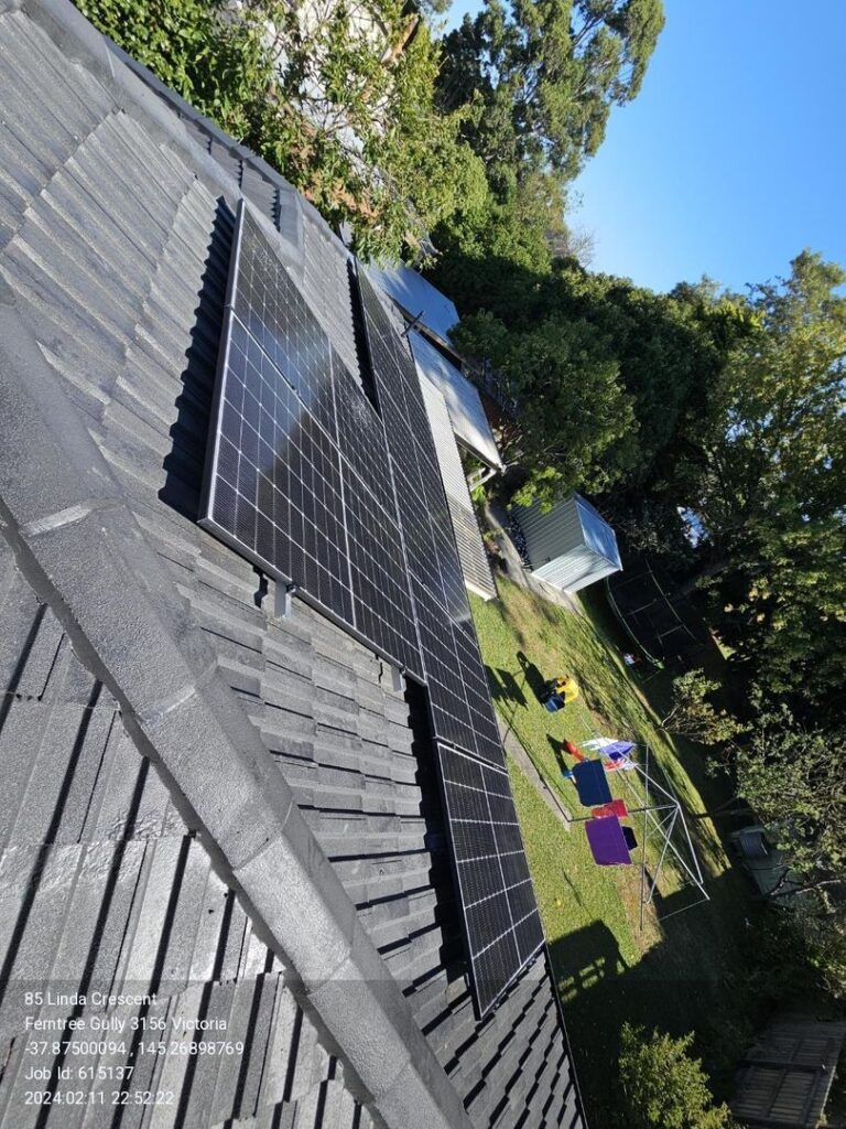 Two rows of solar panels installed on the roof of a house with a backyard featuring trees, a shed, and children's play equipment.