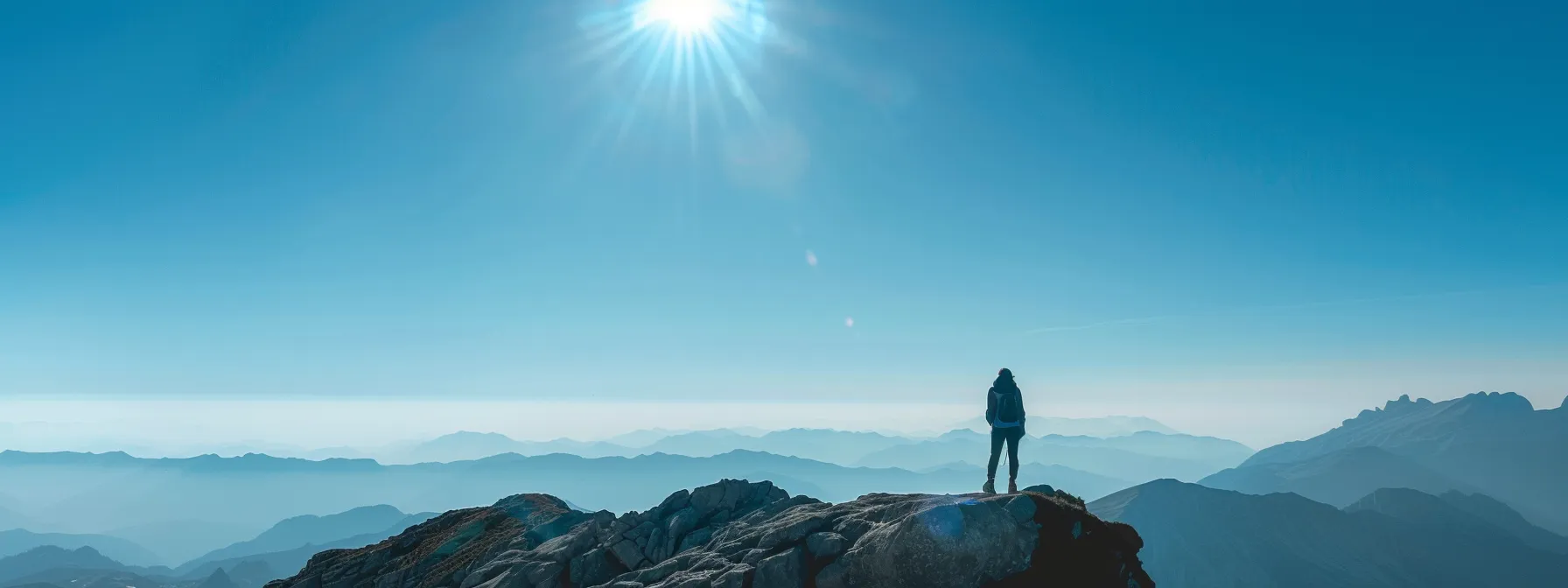 a person standing confidently on top of a mountain, looking out at a clear blue sky, symbolizing overcoming barriers with mindset and success coaching.