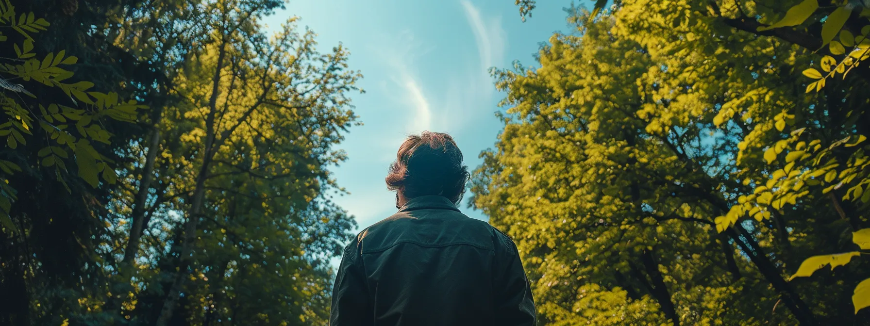 a person standing in a serene forest, surrounded by vibrant green trees and a clear blue sky above, symbolizing emotional balance and healing from ptsd through psych-k therapy.